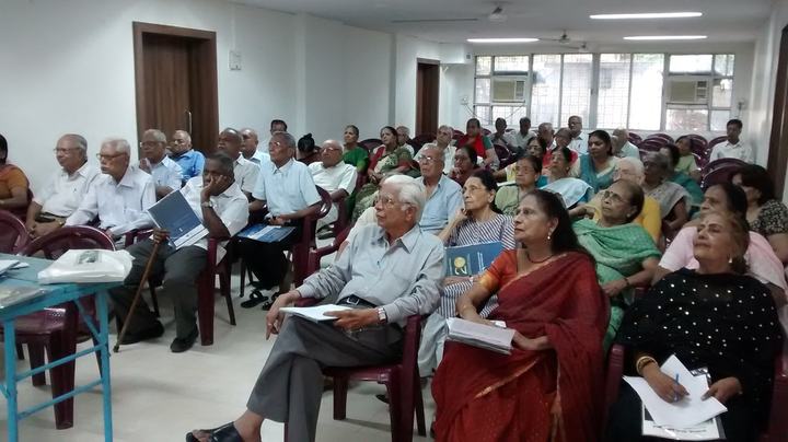 An audience of older adults seated in rows, listening to a talk.