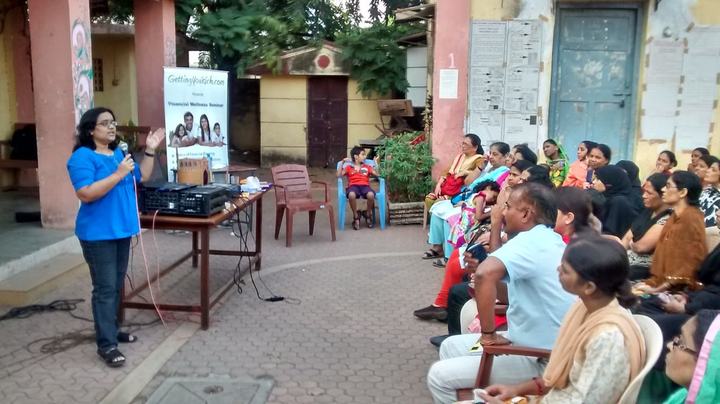 A presenter speaks to an audience seated outdoors in a courtyard.